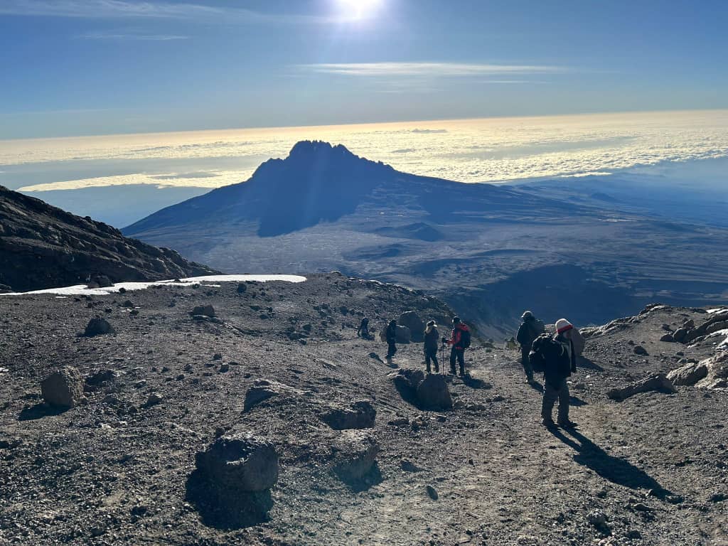 Hikers descending rocky terrain beneath a jagged mountain and bright clouds