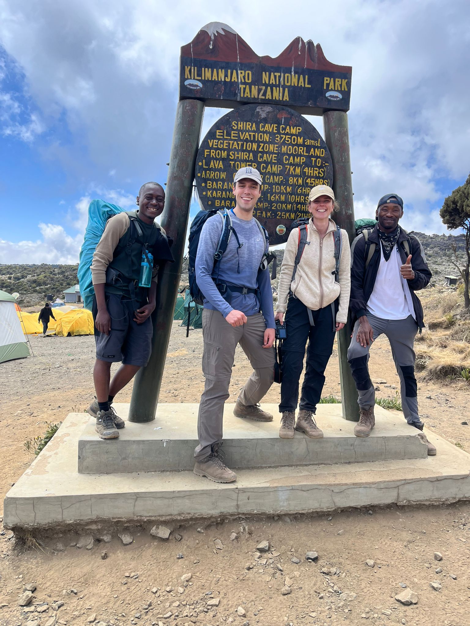 Four climbers gathered at a mountain sign beneath a blue sky
