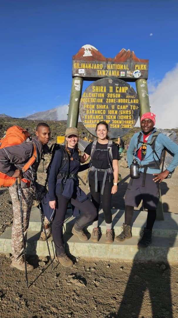 Four hikers posing beside a colourful wooden sign in open mountain terrain