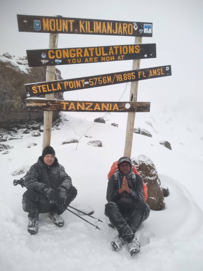 Two climbers sitting in snow beneath a wooden mountain sign