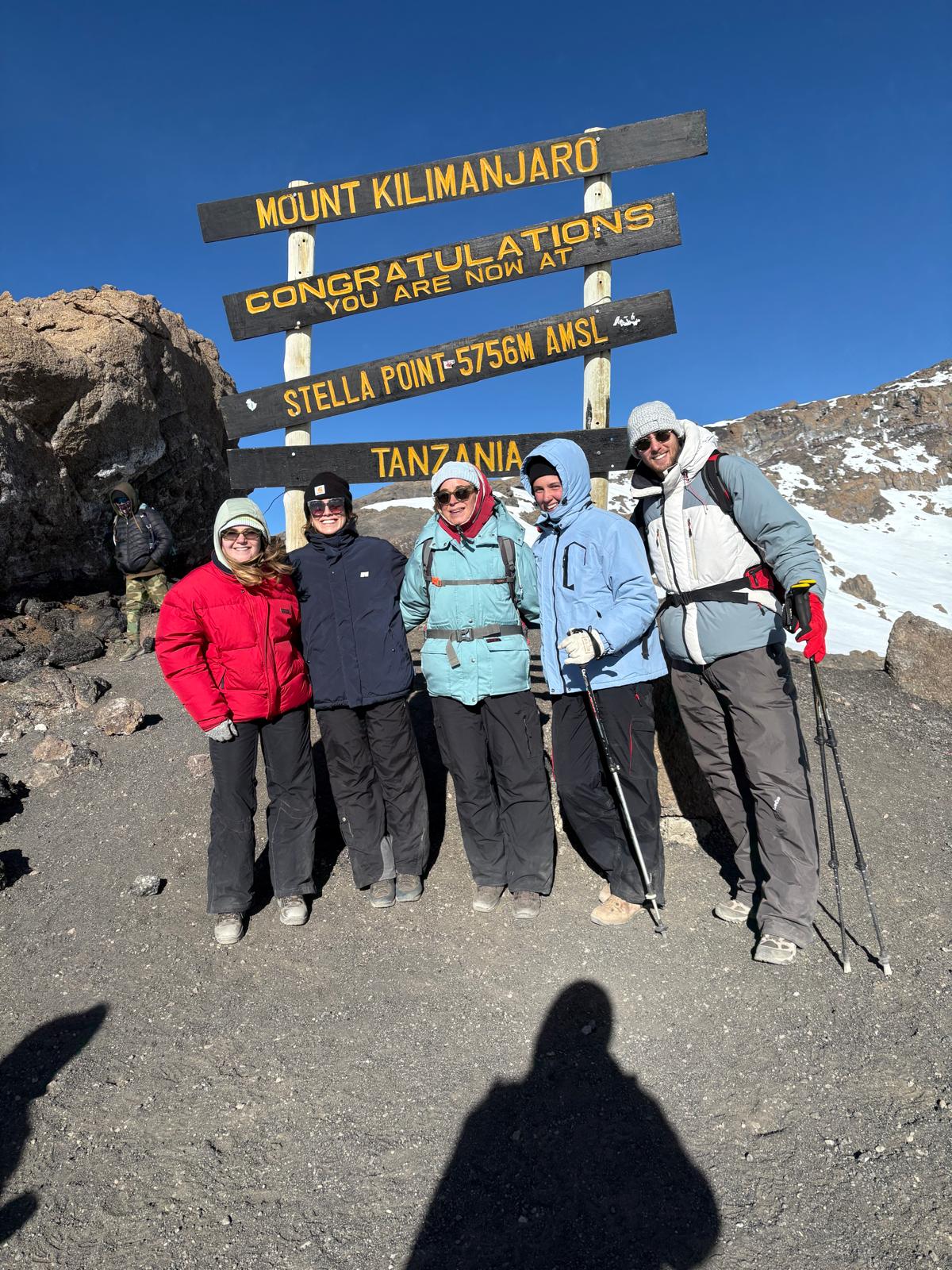 Five climbers posing beside a wooden mountain sign beneath a blue sky