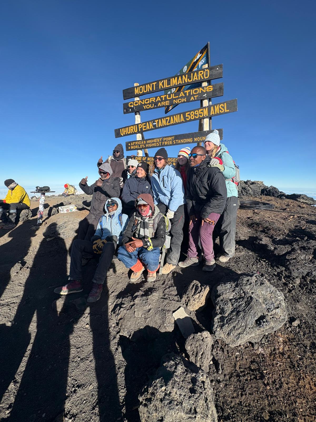 A large group celebrating around wooden summit signs on rocky ground