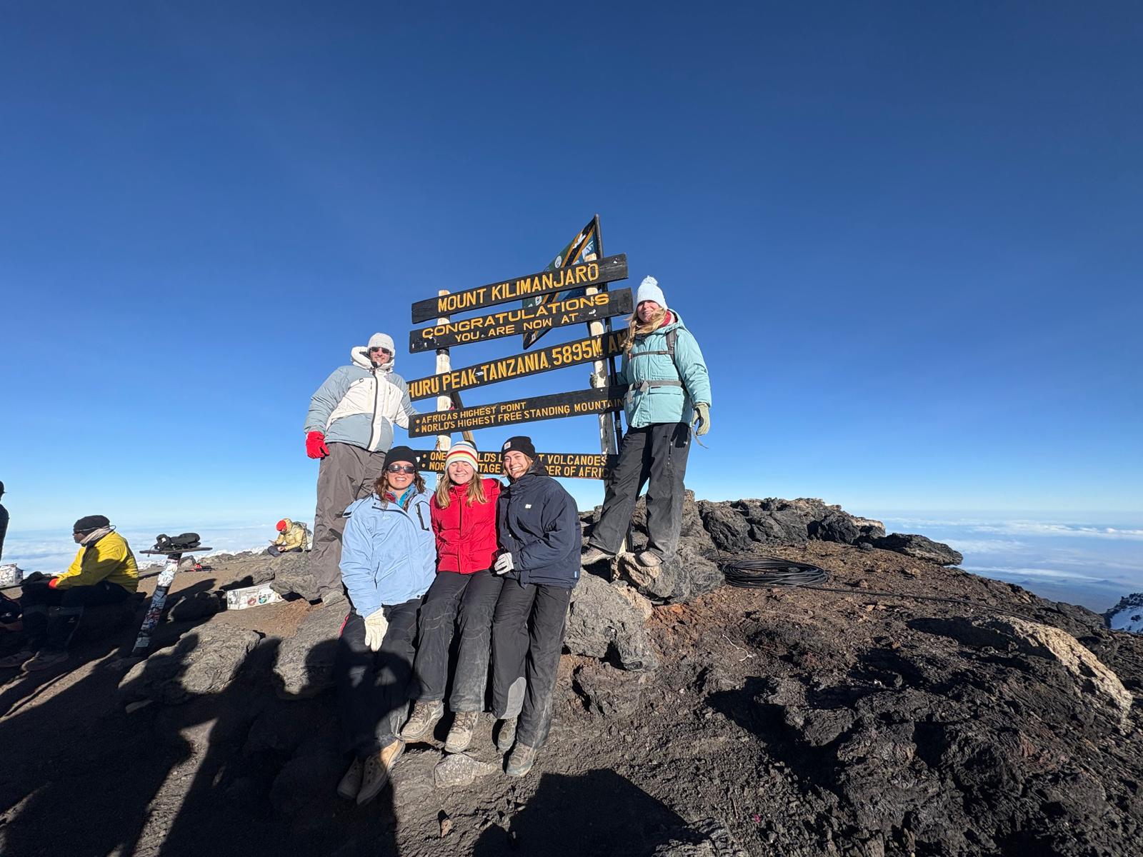 Five climbers standing beside a summit sign on dark volcanic rock