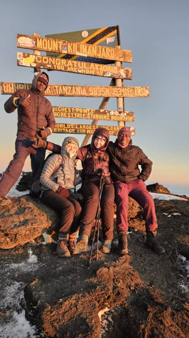 Climbers sitting beside a wooden summit sign in golden light